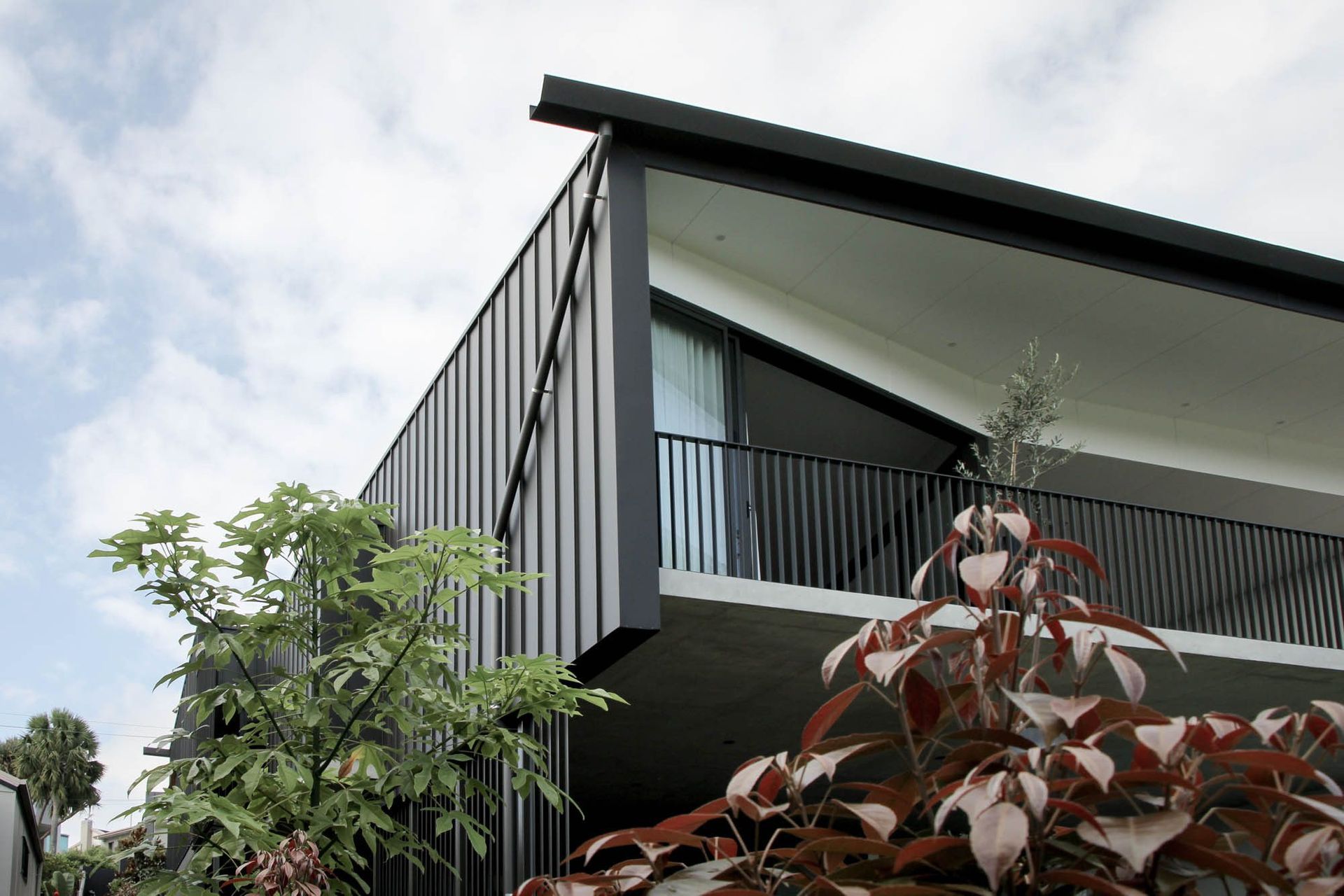 The Dual House - Exterior. Angled roof and balcony above lush planting.