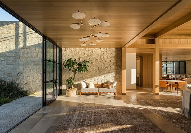 A warm, open-plan interior with a timber-lined ceiling and exposed timber structural framing. Full-height black-framed glazing opens to a paved courtyard, while a textured pale stone wall forms the backdrop to a sofa seating area. The space connects visually to a dining area and features stone or concrete-look flooring, a large neutral rug, and a sculptural multi-arm pendant light.