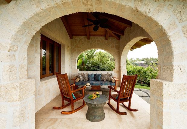 Covered outdoor terrace or veranda with prominent cream stone masonry arches, a warm timber vaulted ceiling, timber-framed window, and tiled or stone flooring. Two wooden rocking chairs face a built-in cushioned bench around a small round table, creating a sheltered seating area overlooking greenery and rooftops. The space has a rustic Mediterranean or tropical resort character.