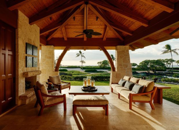 A covered tropical outdoor lounge or lanai with a high exposed timber trussed ceiling, ceiling fan, stone-clad columns and walls, and warm timber-framed seating arranged around a coffee table. The open arched facade looks onto manicured grounds, palm trees and a distant waterfront or ocean view.