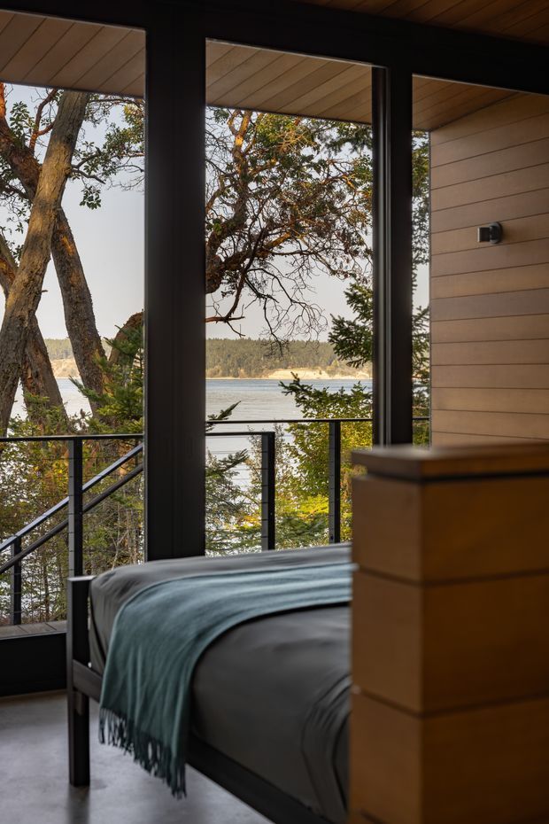 A moody bedroom overlooking a wooded waterfront through large floor-to-ceiling windows. The room has a dark-framed bed with grey bedding and a blue-green throw, a warm timber chest of drawers, a timber-lined ceiling, and horizontal timber wall cladding. Black metal balcony railings and exterior steps are visible beyond the glazing.
