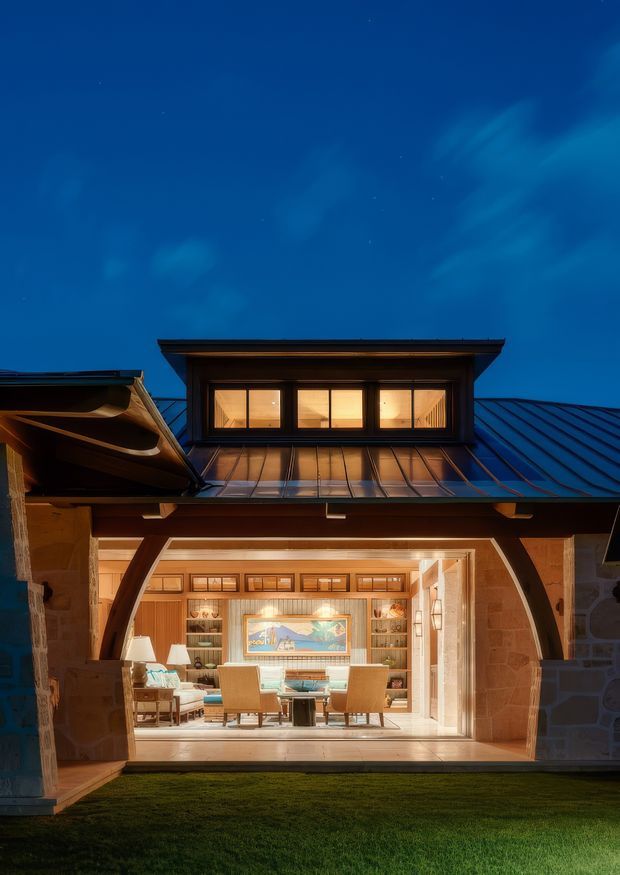 A dusk exterior view of a substantial house with a steep metal roof, projecting dormer windows glowing warmly from within, and a large open arched stone-framed opening revealing an inviting timber-lined living or dining space. The facade combines stone masonry, dark timber framing, and standing-seam metal roofing, with a lawn in the foreground and dramatic deep-blue evening sky.