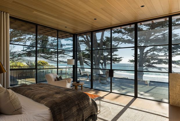 A bright, refined bedroom with a bed in the foreground and a sitting area beside expansive black-framed floor-to-ceiling glazing. The room has a warm timber-lined ceiling, pale flooring, neutral upholstered furniture, and direct views toward a balcony, swimming pool, and coastal landscape. Strong natural light creates graphic shadows across the floor.