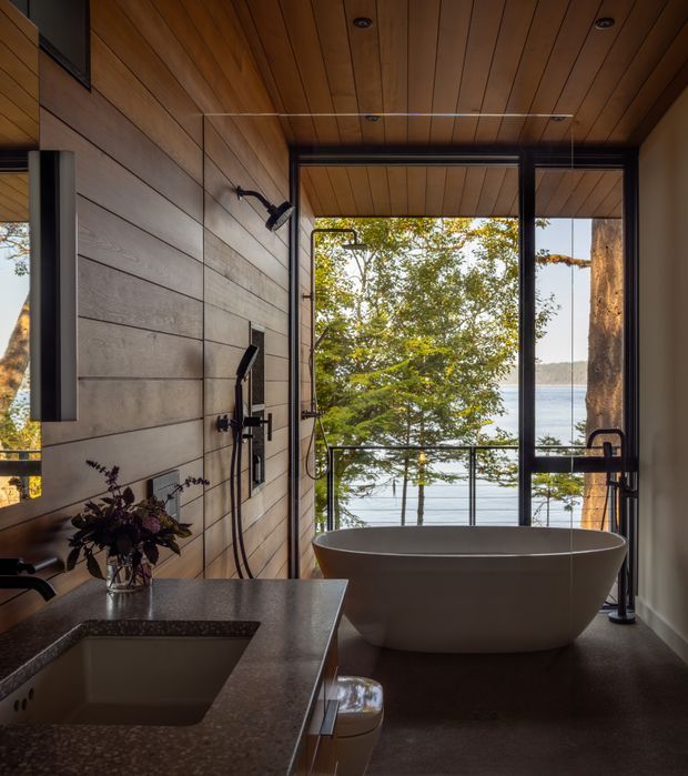 A refined, nature-facing bathroom with a freestanding oval bathtub positioned beside full-height black-framed glazing overlooking a wooded lakeside setting. The room features a timber-lined ceiling, horizontally clad wall panels, a dark stone vanity with integrated basin, exposed shower fittings, recessed ceiling lights, and a minimalist black metal tapware and frame palette.