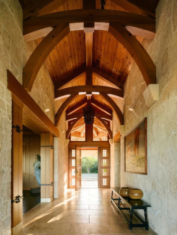 A long, grand entry hall with pale textured stone walls, warm timber panelled vaulted ceiling, and exposed heavy timber arches and beams. Large timber-and-glass double doors open to the exterior at the far end, while a timber side door reveals a small adjoining space. The hall has a pale stone tile floor, black metal console table, framed artwork, decorative vessels, and a dramatic hanging lantern.