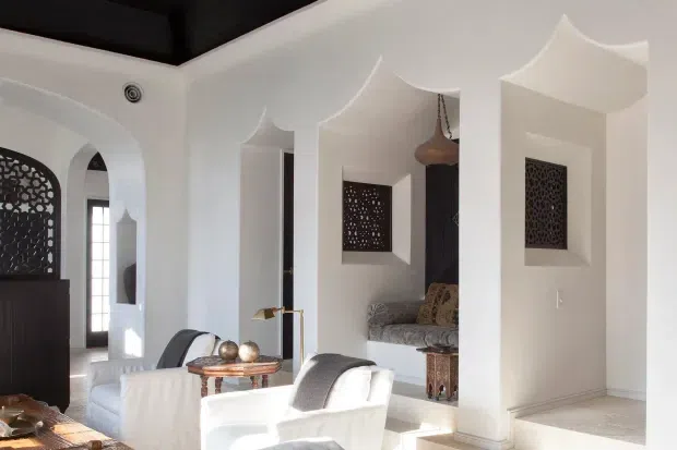 Bright, refined living room with white sculptural arched wall niches and pointed, scalloped ceiling transitions. The room includes white upholstered lounge chairs, a small round wooden table, patterned dark decorative screens set into the niches, and dark timber architectural accents. An arched opening leads toward an adjacent hallway or room.