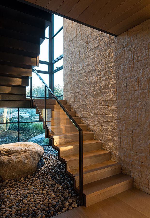 Interior architectural photograph of a sculptural timber staircase with dark metal stringers and a slim black handrail. The stair rises beside a tall glazed wall, bringing views of landscaped grounds indoors. A rough-faced masonry feature wall contrasts with the warm timber ceiling and stair treads, while a pebble-filled floor with a large natural boulder creates an organic Japanese-inspired garden detail beneath the stairs.