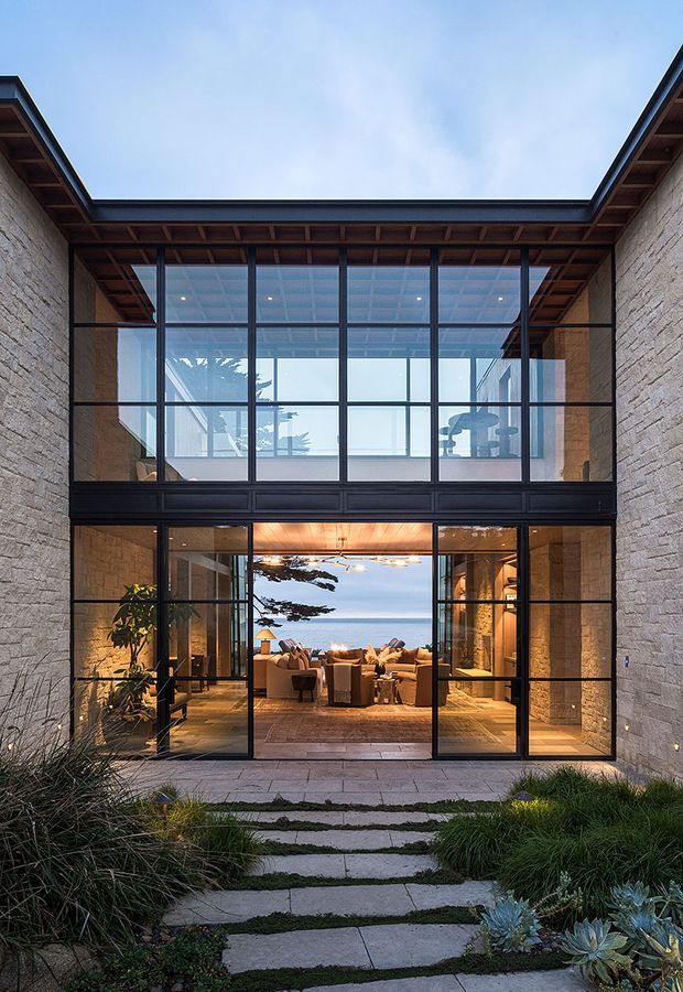 Exterior view of a contemporary two-storey courtyard-facing residence with pale stone walls, expansive black-framed glazing, and a recessed timber-lined roof canopy. A landscaped stepping-stone path leads to open glazed doors, revealing a warmly lit living room and a second-level interior. The image also shows a coastal ocean outlook through the house, low-maintenance planting, and strong indoor-outdoor connection.