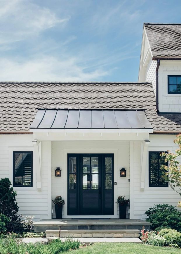 Front exterior view of a refined white-clad house with a prominent gabled upper volume, grey shingle roofing, dark-framed windows and glazed double entry doors. The covered entrance features a standing-seam metal canopy, white square columns, wall-mounted lanterns, and landscaped planting around stone steps.
