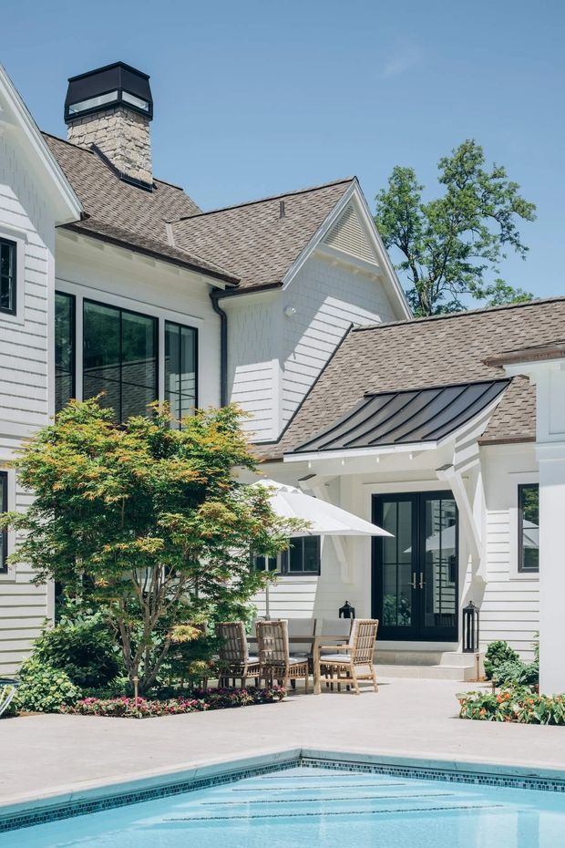 Exterior view of a bright, multi-gabled white residential home with shingle siding, a stone chimney, pitched shingle roofs, dark-framed glazed doors and windows, and a covered patio dining area beside a swimming pool. Mature landscaping and planting surround the paved courtyard.
