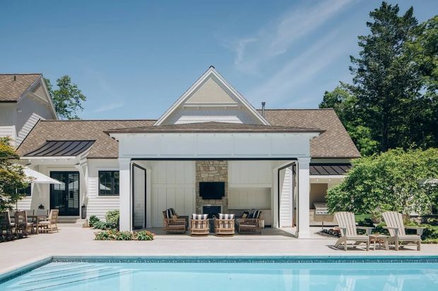 A bright rear exterior view of a large traditional-style home beside a swimming pool. The white-painted facade has multiple gabled roof sections, shingle-style roofing, dark metal roof accents, and an open pool cabana or outdoor living pavilion with a stone fireplace, seating, and wide folding doors. A paved terrace, outdoor dining area, garden planting, and pool occupy the foreground.