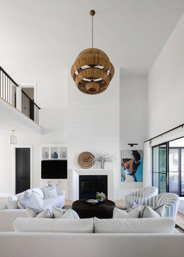 Bright, airy double-height living room with a white shiplap-style feature wall, central fireplace, large woven pendant light, black-framed glazing, and an upper-level balcony or landing with black balustrade. The room is furnished with white sofas, striped armchairs, a dark round ottoman coffee table, patterned cushions, and restrained coastal artwork.