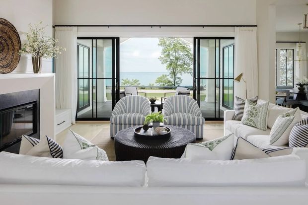Bright, airy living room arranged around a large white sectional sofa, two striped armchairs, and a dark round coffee table. Floor-to-ceiling black-framed glazed doors open toward a balcony or terrace with a visible water view. The room includes a built-in fireplace, light walls, pale flooring, white curtains, and coastal-inspired furnishings and accents.