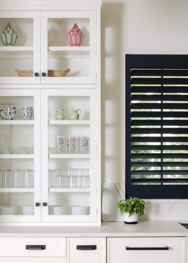 Bright, close-up view of a white kitchen with tall glass-front display cabinetry, open shelves holding decorative ceramics and glassware, a pale stone benchtop, white drawers with dark bar handles, and a black horizontal-slat window blind. A small potted plant sits on the benchtop beside the window.