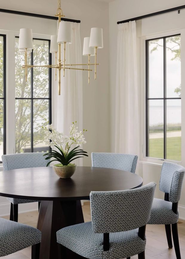 Bright, elegant dining room with a large round dark wood dining table surrounded by patterned upholstered chairs. Tall black-framed windows, white walls, sheer floor-to-ceiling curtains, and a sculptural brass multi-arm chandelier create a refined, airy interior. A floral arrangement sits at the centre of the table.