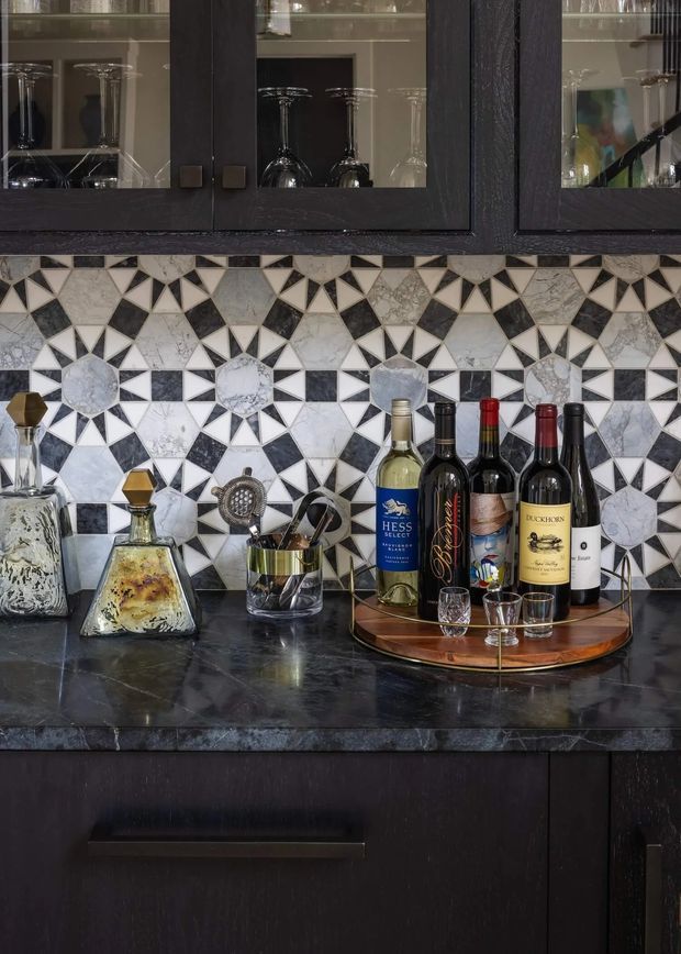 Close-up of a sophisticated built-in home bar, featuring dark timber cabinetry with glass-front upper doors, a polished dark stone benchtop, and an ornate geometric mosaic tile splashback in white, grey, and black. Wine bottles, decanters, cocktail tools, and glasses are arranged on a round wooden tray and across the counter.