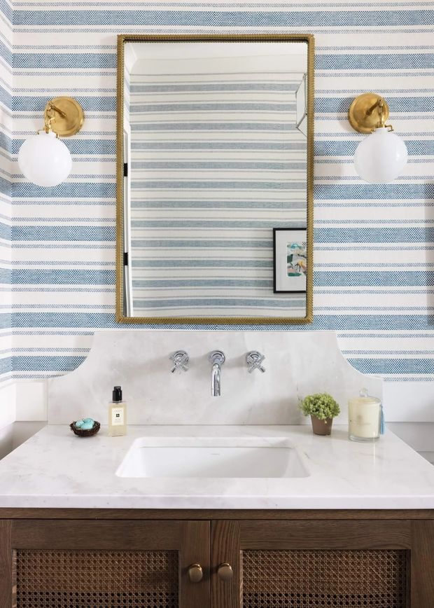 A stylish bathroom vanity with a white marble or stone countertop, integrated rectangular basin, exposed chrome cross-handle tapware, and dark timber cabinetry with woven cane-front doors. The wall is covered in blue-and-white horizontal striped wallpaper or patterned wall finish. A large brass-framed rectangular mirror is flanked by brass wall sconces with white globe shades. Small toiletries, a plant, and a candle sit on the vanity.