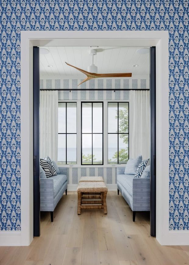 A bright, narrow sitting room viewed through a wide doorway. The room features two pale blue upholstered sofas facing each other, a woven rattan ottoman, tall black-framed windows with sheer white curtains, patterned blue-and-white wallpaper, striped wall panels beneath the windows, a white paneled ceiling, and a sculptural wood ceiling fan. Pale timber flooring and coastal-inspired blue, white, and natural textures create an airy, refined interior.