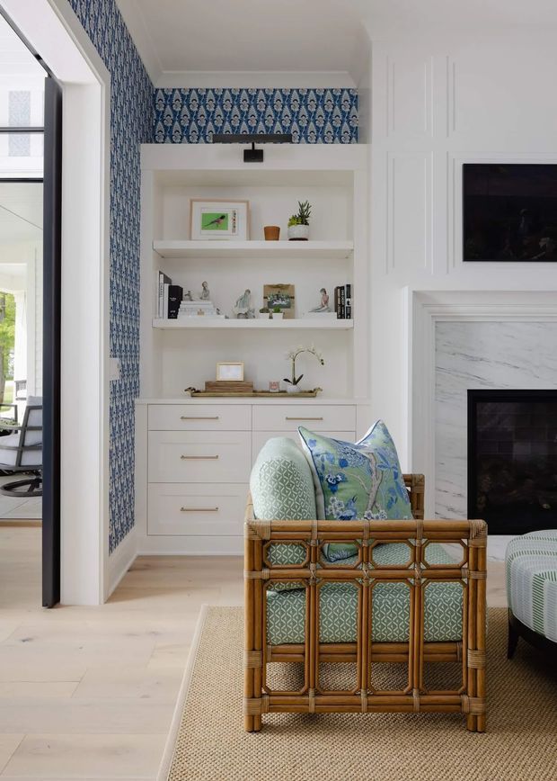 Bright, refined living room with a built-in white shelving and drawer unit, patterned blue-and-white wallpaper, a paneled fireplace surround with television above, and a woven rattan armchair upholstered in blue and pale green patterned fabric. Pale timber flooring, a natural woven rug, and white architectural detailing create a polished, traditional coastal feel. An adjacent outdoor area is visible through the open doorway.