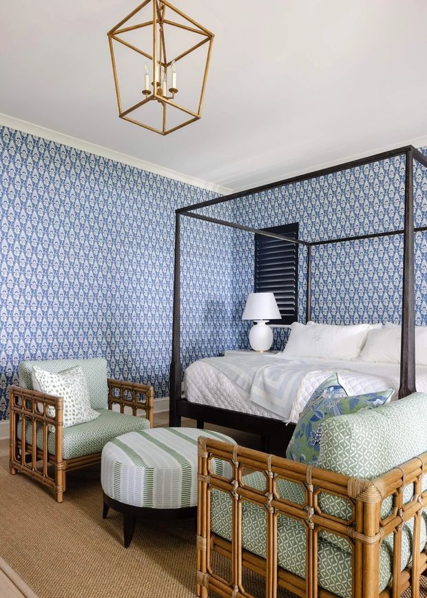 A traditionally styled bedroom with a dark four-poster bed, white bedding, patterned blue-and-white wallpaper, and woven rattan seating. Two rattan armchairs and a round upholstered ottoman sit on a natural-textured carpet. A geometric brass pendant light hangs from the white ceiling, while dark plantation shutters provide contrast behind the bed.