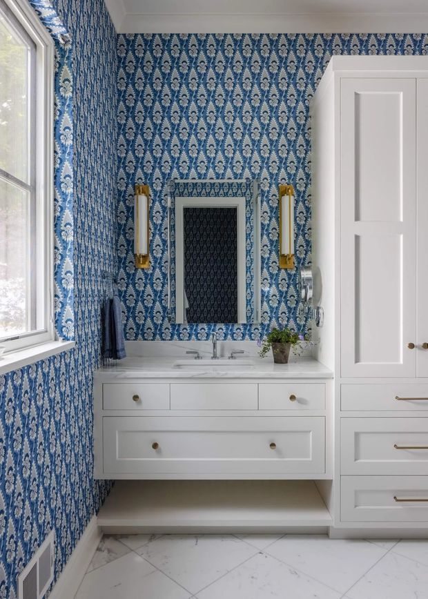 A refined bathroom with a floating white vanity, integrated double-sink basin, brass fixtures and wall sconces, framed mirror, and tall built-in storage cabinetry. The walls are covered in a bold blue-and-white patterned wallpaper, while the floor is finished in pale marble-look tiles. A large window provides natural light, and traditional panelled cabinetry and trim create a classic, decorative character.
