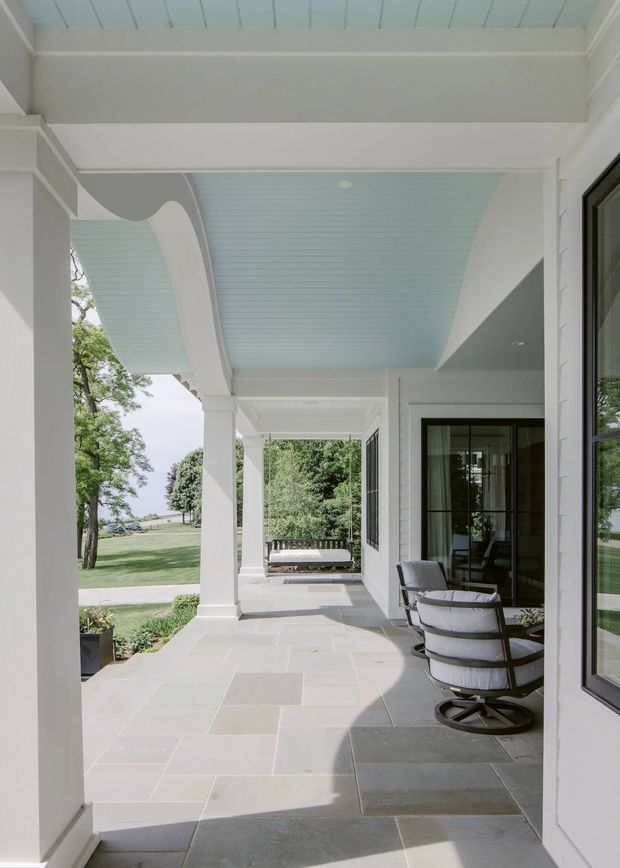 A bright, covered outdoor veranda or porch adjoining a white-clad home. The space features substantial white square columns, a pale blue beadboard-style ceiling with curved detailing, large dark-framed glazed doors and windows, and a light stone-paved floor. Outdoor lounge chairs and a bench sit along the veranda, which opens onto a landscaped lawn and garden.