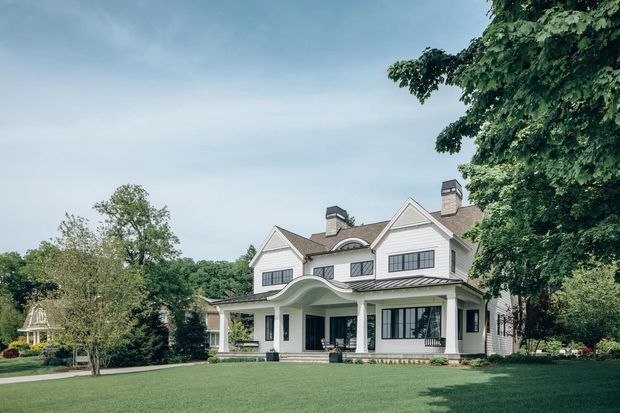 A large white multi-storey residential house viewed from the front across an expansive lawn. The facade features gabled roof sections, multiple chimneys, dark-framed windows, a covered entry porch, and a distinctive curved canopy over the main entrance, surrounded by mature trees and landscaped grounds.