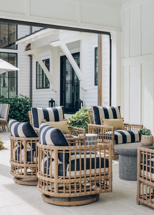 A bright, refined outdoor lounge courtyard or covered patio furnished with several woven rattan armchairs and ottomans upholstered in bold navy-and-white stripes. A small central table with greenery sits among the seating. White horizontal weatherboard walls, black-framed glazing and doors, and white structural beams form the backdrop, with potted plants adding an organic touch. The space has a relaxed coastal character and a strong indoor-outdoor connection.