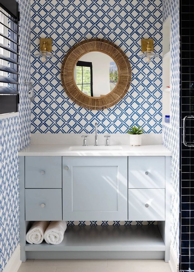A bright, compact bathroom vanity area with a white floating-style cabinet, integrated white basin and chrome tapware. The walls are clad in a bold blue-and-white geometric patterned tile, complemented by a round woven-framed mirror and brass wall sconces. Open lower shelving holds rolled towels, and a small potted plant adds greenery. Black-framed louvred glazing and a black tiled shower enclosure are visible at the sides.