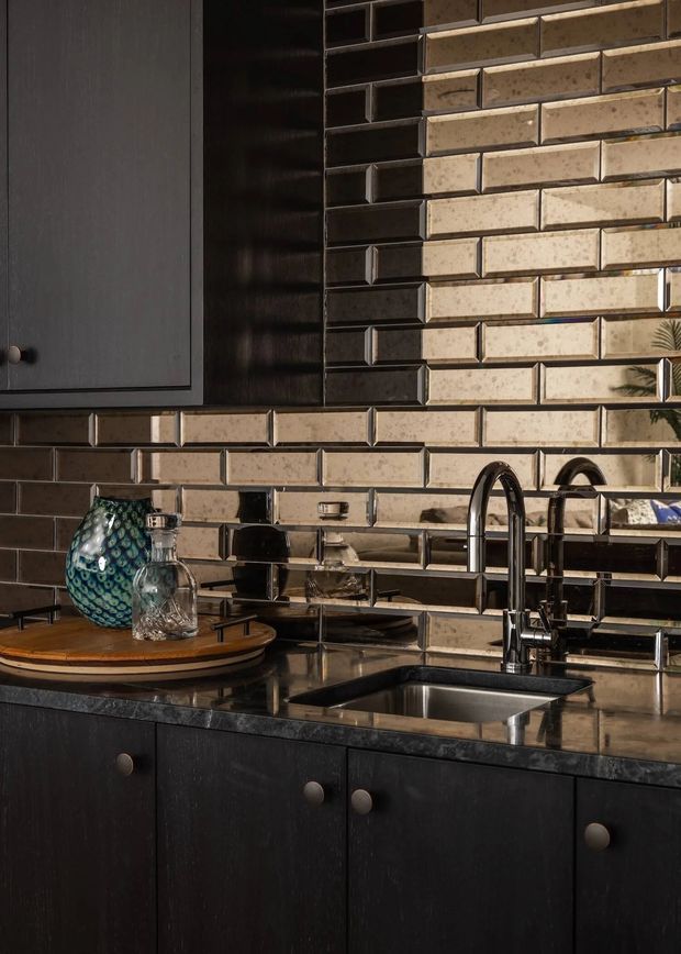 Close-up view of a dark, sophisticated kitchen with black wood cabinetry, a polished dark stone benchtop, stainless-steel double sink and curved chrome tapware. The backsplash is composed of glossy, elongated beige and charcoal brick tiles with contrasting grout, creating a strong textured feature wall. Decorative glassware and a patterned vase sit on a round timber tray.