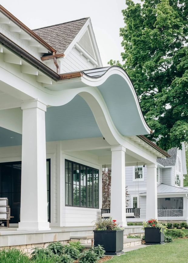 Exterior view of a large white weatherboard house with prominent covered porch, classical square columns, black-framed windows, gabled roof sections, copper-toned gutters, and a distinctive undulating curved porch canopy. The landscaped front garden and mature tree frame the facade.
