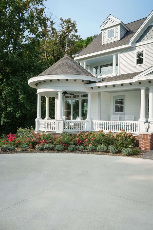 Exterior view of a large white clapboard house with a prominent rounded veranda, classical columns, balustrades, multiple gables, shingle roofing, and a landscaped garden along a broad concrete driveway. The facade combines traditional detailing with a distinctive conical-roofed porch.
