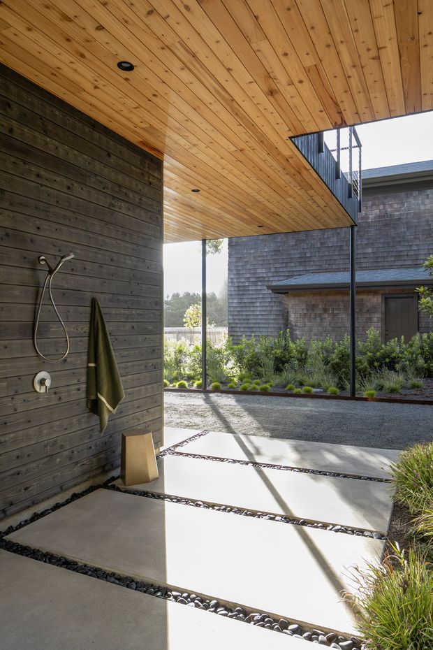 Covered outdoor shower courtyard with dark horizontal timber wall cladding, a warm timber-lined soffit, minimalist metal supports, and large pale concrete paving slabs separated by narrow bands of dark river stones. The view opens onto a landscaped garden and a shingle-clad building, creating a clean, contemporary indoor-outdoor transition.