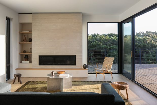 A contemporary living room with a large pale stone or timber-look fireplace feature wall, integrated linear fireplace, and open shelving. Floor-to-ceiling black-framed glazing opens to a timber deck and leafy outdoor setting. The room includes a dark sectional sofa, a round metallic coffee table, a patterned area rug, and a warm-toned lounge chair, creating a refined indoor-outdoor connection.