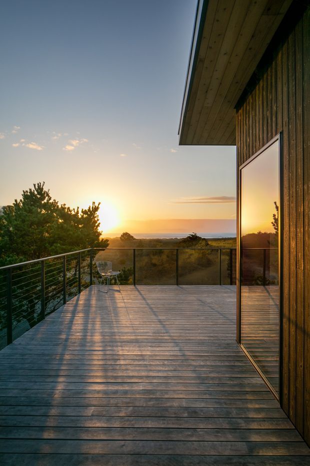 A timber-clad modern dwelling with a spacious elevated deck overlooking a coastal or ocean-facing landscape at sunset. The deck has wide timber floorboards, slim dark metal balustrades with transparent panels, and large reflective glazing integrated into the dark vertical timber facade. A single wire-frame chair provides scale, while warm low-angle light creates long shadows across the deck.