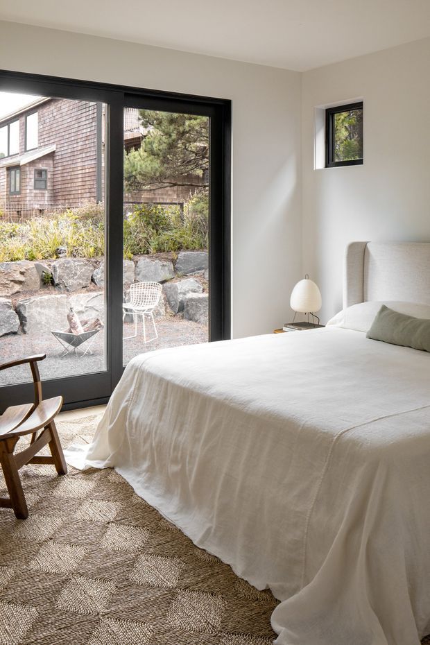 A bright, calm bedroom with a large white upholstered bed dressed in white linen, a small rounded bedside lamp, and a sculptural wooden lounge chair. Black-framed floor-to-ceiling glazing opens toward a landscaped rocky courtyard, while a small square window adds natural light. The room has warm neutral finishes and a textured natural-fibre rug.