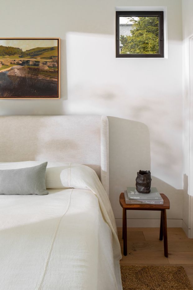 A softly lit bedroom corner featuring a cream upholstered bed with a curved wingback headboard, layered white and pale grey bedding, and a dark timber bedside table holding books and a small sculptural vessel. A framed landscape artwork hangs on the pale wall beside a small dark-framed window, with light timber flooring and a woven rug completing the calm, neutral interior.