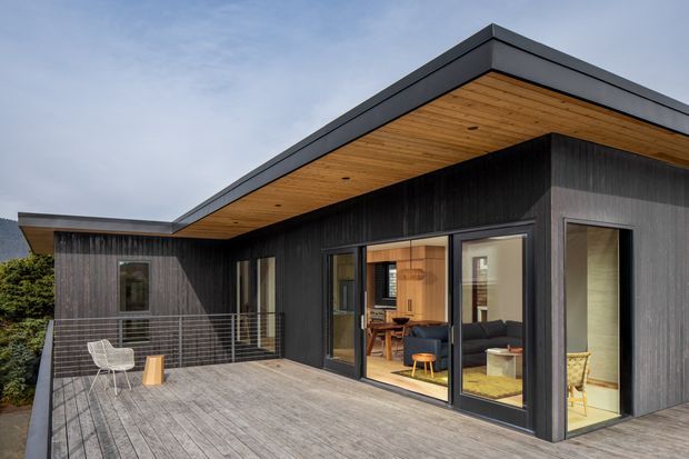 Exterior view of a contemporary single-storey home with a strong flat-roofed, L-shaped form. Dark vertical timber cladding contrasts with warm timber soffits and a broad grey timber deck. Large black-framed glazed sliding doors open onto the deck, revealing a living area and adjacent interior spaces. The design has clean horizontal lines, recessed roof edges, and a minimalist outdoor railing.