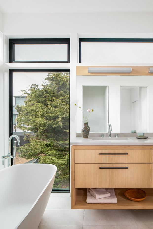 Bright contemporary bathroom with a freestanding white bathtub beside tall black-framed windows, a wall-mounted light timber vanity with open shelving, white countertop and integrated basin, twin mirrors or recessed mirror panels, and brushed metal tapware. Large windows overlook trees and bring in abundant natural light.