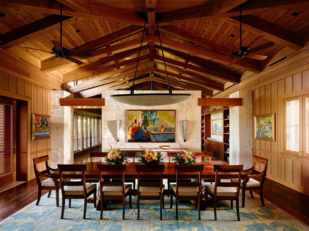 A warm, richly timber-lined dining room with a long rectangular dining table surrounded by wooden chairs. The room features a dramatic exposed timber vaulted ceiling with heavy beams, ceiling fans, pendant lighting, timber wall panelling, artwork, woven window blinds, and a textured stone or plaster fireplace feature wall. The palette is dominated by honey-toned wood, deep brown furniture, and muted blue-green patterned flooring or rug.