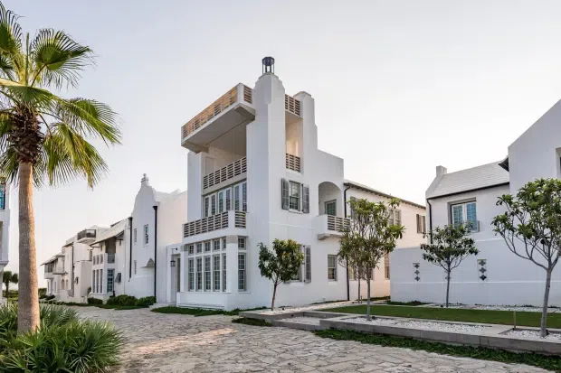 Exterior view of a white multi-storey residential development with strong Mediterranean and coastal influences. The buildings feature white stucco walls, arched openings, balconies with horizontal metal or timber-look screens, tall glazed doors and windows, pitched and flat roof forms, and a paved pedestrian forecourt with palm and ornamental trees.