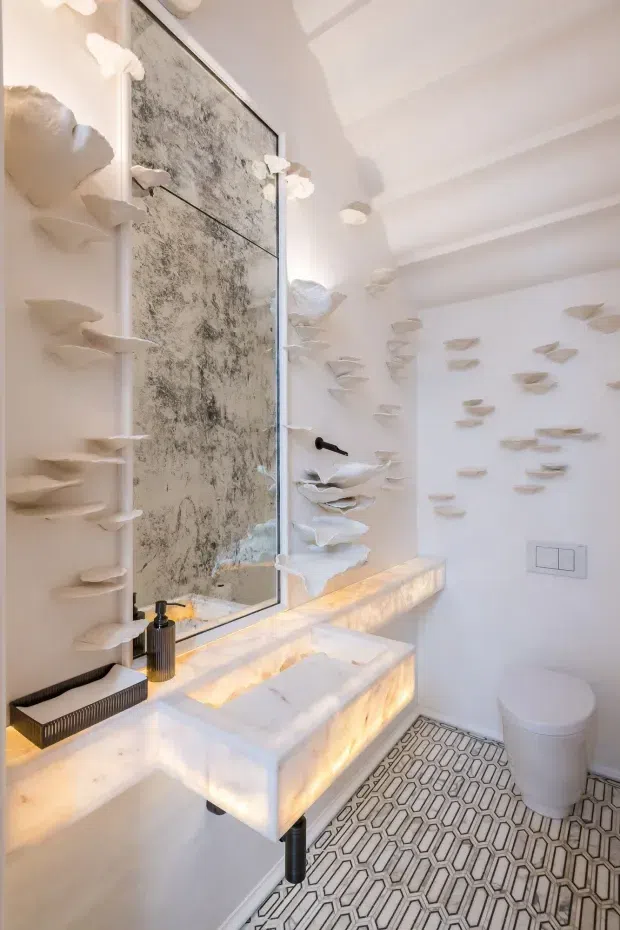 A bright, highly designed bathroom with a floating sculptural vanity and integrated illuminated stone basin. The walls and mirror are decorated with layered white sculptural forms resembling organic shells or rock ledges, while a large mottled feature panel sits behind the mirror. The room has white walls and ceiling mouldings, a wall-mounted tap, a white toilet, and patterned geometric floor tiles in grey and white. Warm concealed lighting beneath the vanity adds a luxurious glow.
