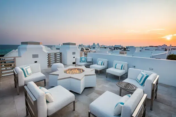 A spacious rooftop terrace overlooking a coastal town and ocean at sunset. The terrace features white and pale grey outdoor lounge seating arranged around a geometric built-in fire pit, with turquoise accent cushions and a small slatted side table. White parapet walls and flat-roofed buildings reinforce the bright coastal setting.