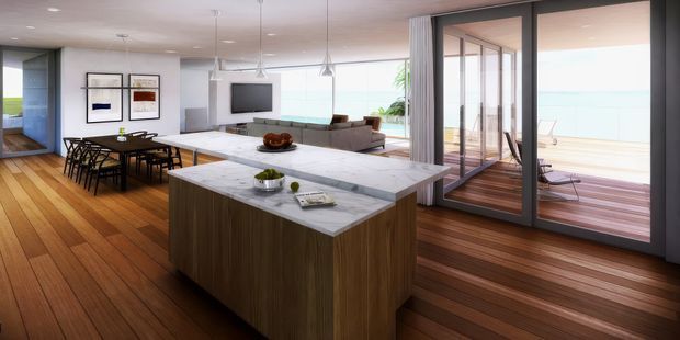 Open-plan contemporary kitchen, dining and living area with a large marble-look island, dark timber cabinetry, warm timber flooring, and extensive glass doors opening onto a timber deck with a visible ocean or water outlook. The space is bright and airy, with a neutral palette and strong indoor-outdoor connection.