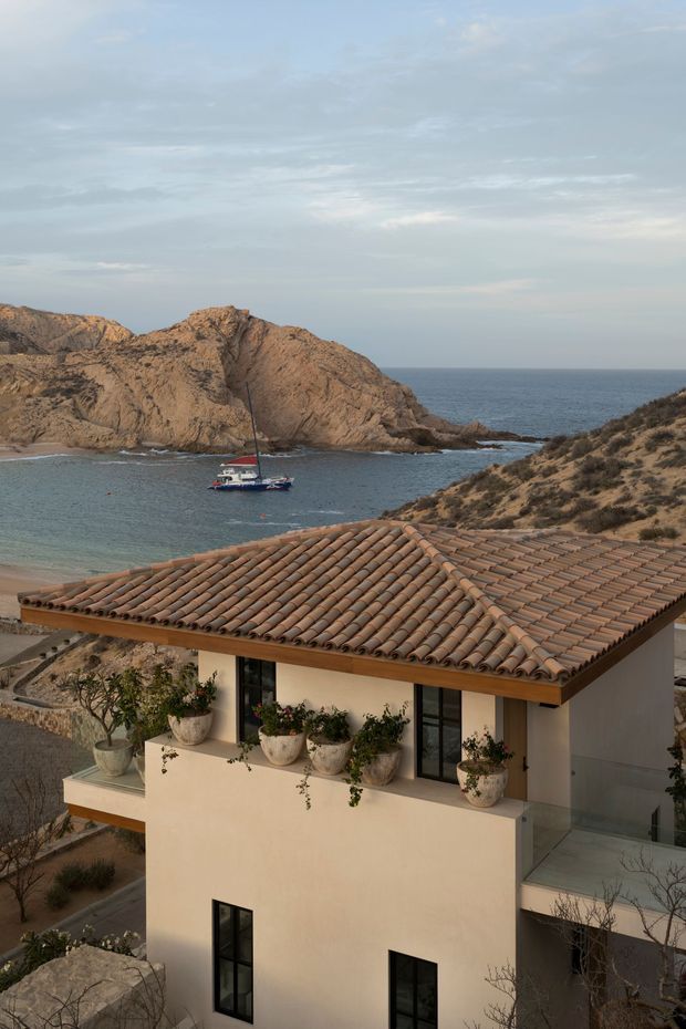 A modern coastal house viewed from above, with a warm terracotta barrel-tile roof, pale rendered walls, dark-framed windows, and a planted terrace. Behind it is a rocky coastal cove with clear blue water and a sailboat, creating a Mediterranean seaside setting.