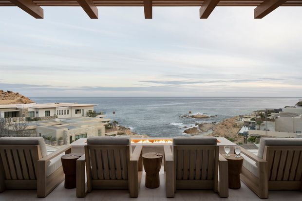 A covered outdoor terrace overlooking a rocky ocean coastline, furnished with a row of timber-framed lounge chairs and small stump-style side tables. A linear fire feature appears along the terrace edge, while a timber pergola provides overhead shade. The scene has a refined coastal resort character with expansive sea views and low-rise buildings on the surrounding rocky shore.