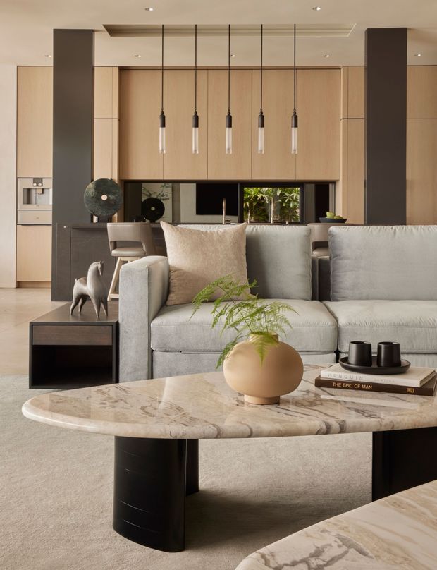 A refined open-plan living room with a contemporary kitchen visible behind it. The foreground features a large curved stone coffee table with dark sculptural supports, a pale upholstered sectional sofa, a small dark side table, a ceramic vase with greenery, and coffee-table books. The kitchen has warm timber cabinetry, dark stone or dark-finished island cabinetry, a black-framed splashback/window, and a row of slim pendant lights. The space uses recessed ceiling lighting and a restrained palette of warm timber, beige, cream, grey, and charcoal.