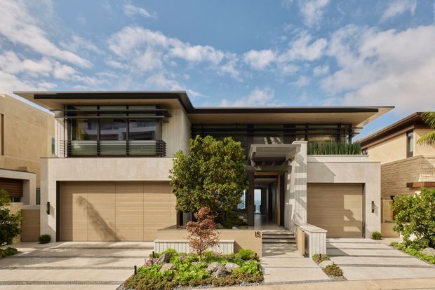 Front exterior view of a contemporary multi-level residence with broad flat roof planes, warm beige rendered walls, large glazed openings with dark frames, integrated double garages, and a recessed entry approached by steps. Low-maintenance landscaped planting and paved driveways frame the facade under a bright sky.