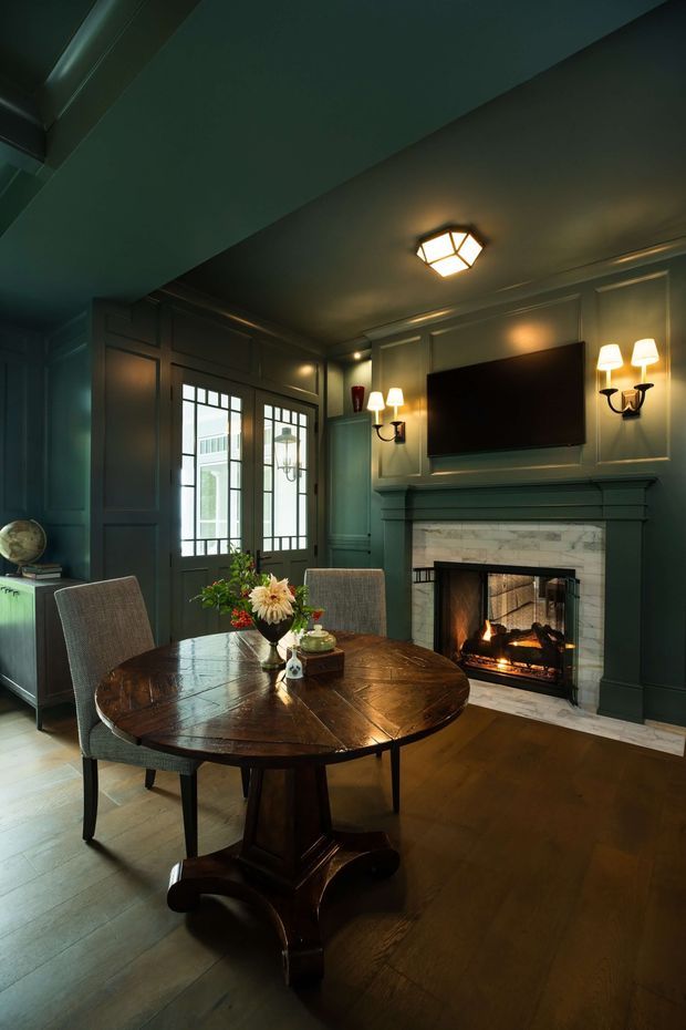 A moody, richly detailed dining room with deep green painted walls and ceiling, dark timber panel mouldings, a round polished wood pedestal dining table, upholstered dining chairs, and a central fireplace with a pale stone or marble surround. A wall-mounted television is integrated above the mantel, with symmetrical wall sconces and glazed double doors providing daylight. The room has an elegant, traditional character with warm firelight and layered decorative lighting.