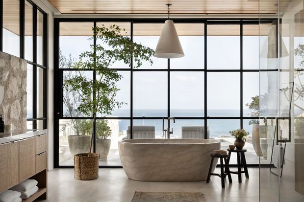 A serene, contemporary bathroom with a freestanding sculptural stone bathtub positioned in front of expansive black-framed glazing overlooking the ocean. The space features a timber-slatted ceiling, pale stone or plaster flooring, a floating timber vanity with a natural stone backsplash, a large pendant light, indoor plants, and a compact stool. The room is bright, refined, and strongly connected to the coastal outlook.